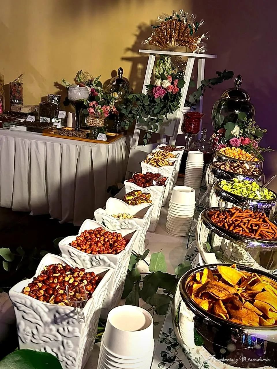 Mesa de snacks completa con jarrones plateados de botanas, nueces y enchilados